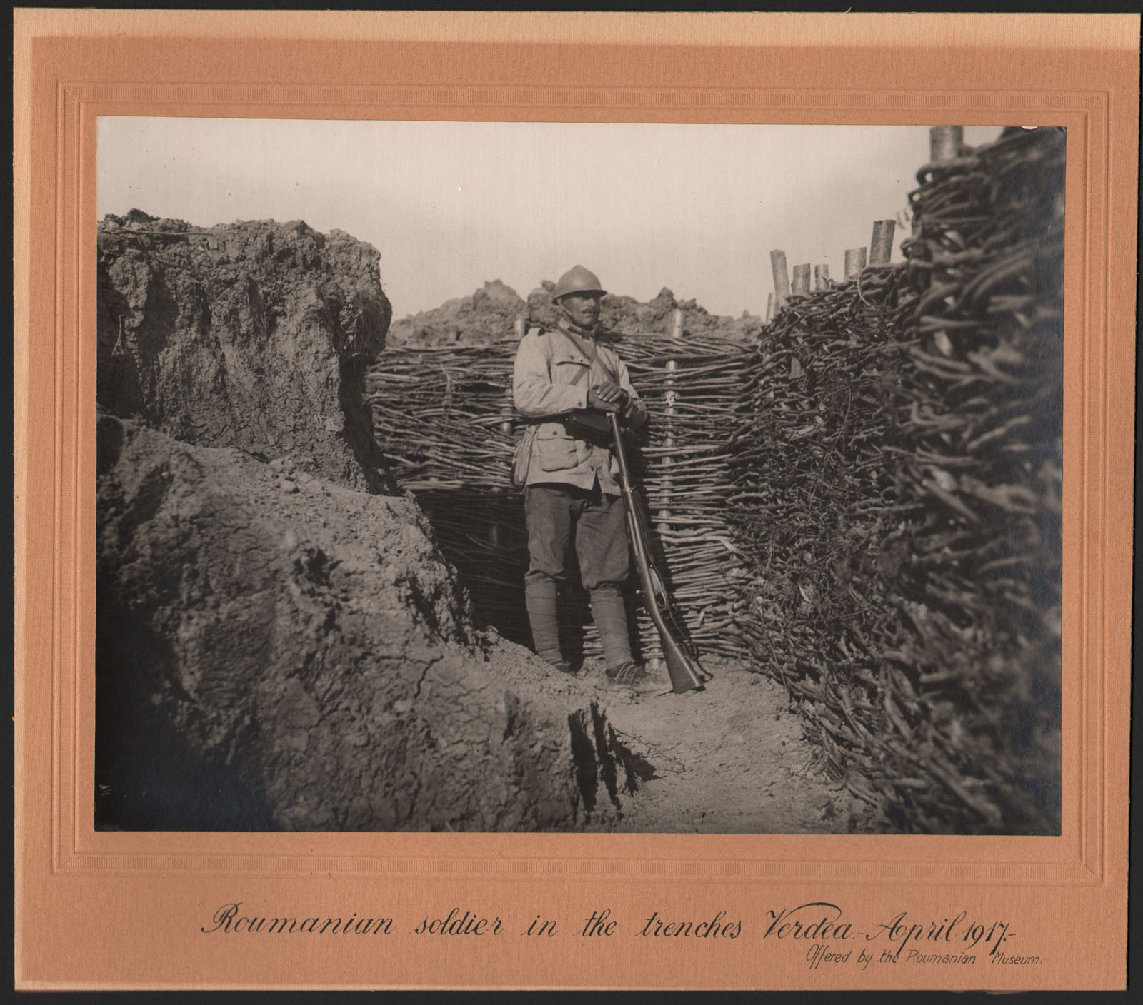 Black and white photograph of a soldier in uniform holding a broom, standing in a trench.