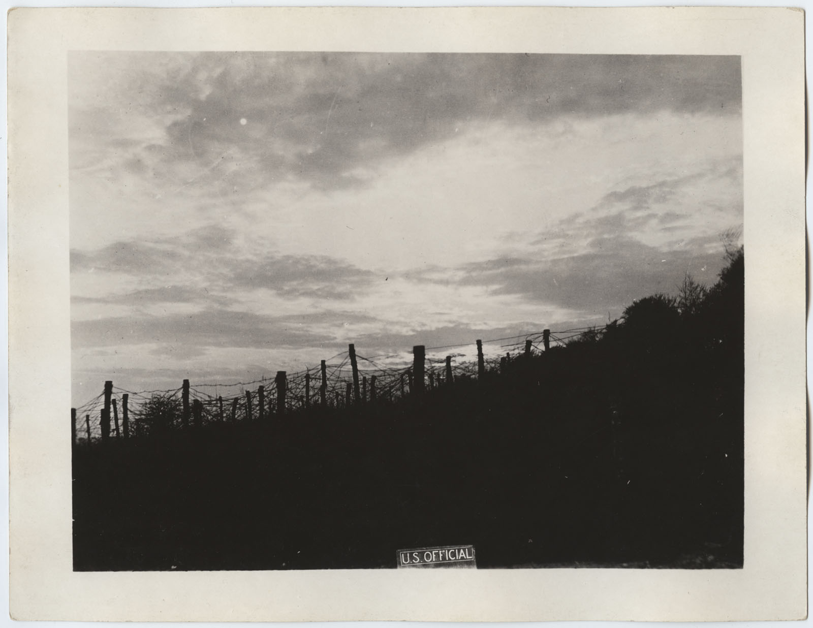 Black and white photograph. A hillside crowned with barbed wire fencing, silhouetted against the sky