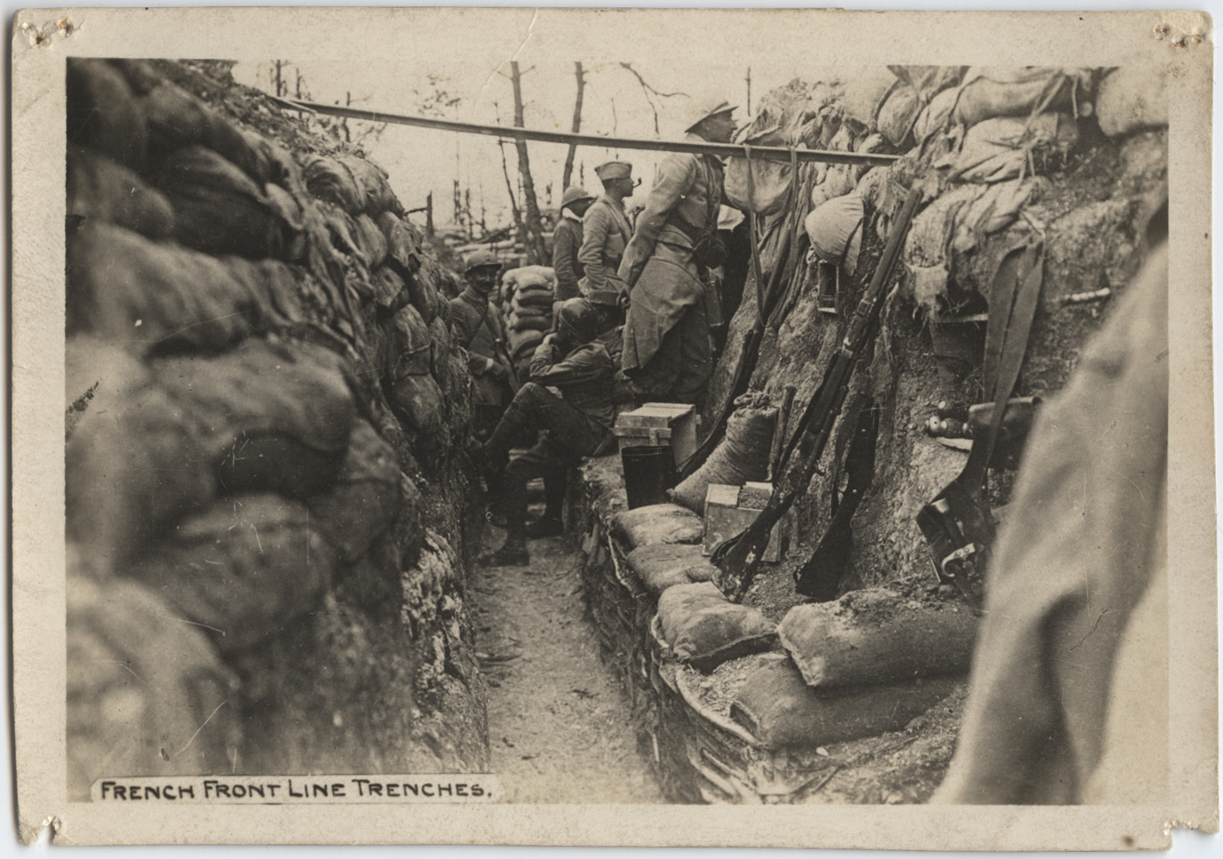 Black and white photograph. Several soldiers in uniform look out over the edge of a trench lined with sandbags