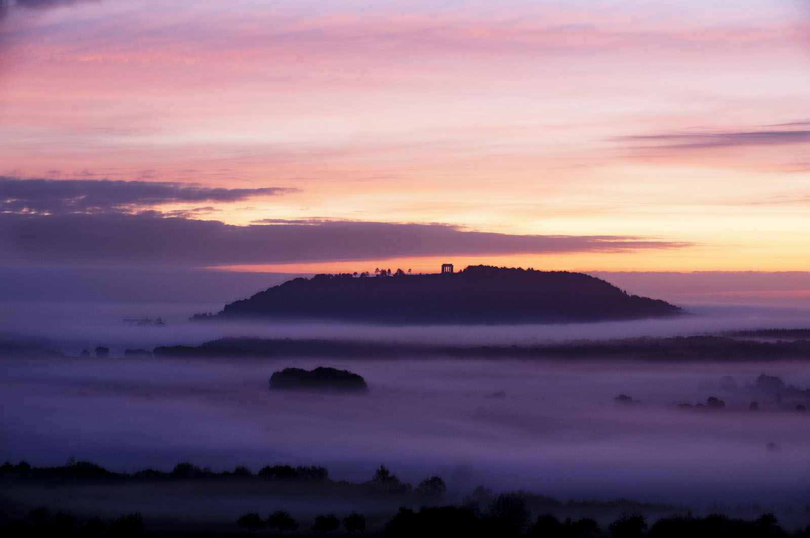 Modern photograph. A hill rises out of foggy fields, lit purple and orange by sunrise.
