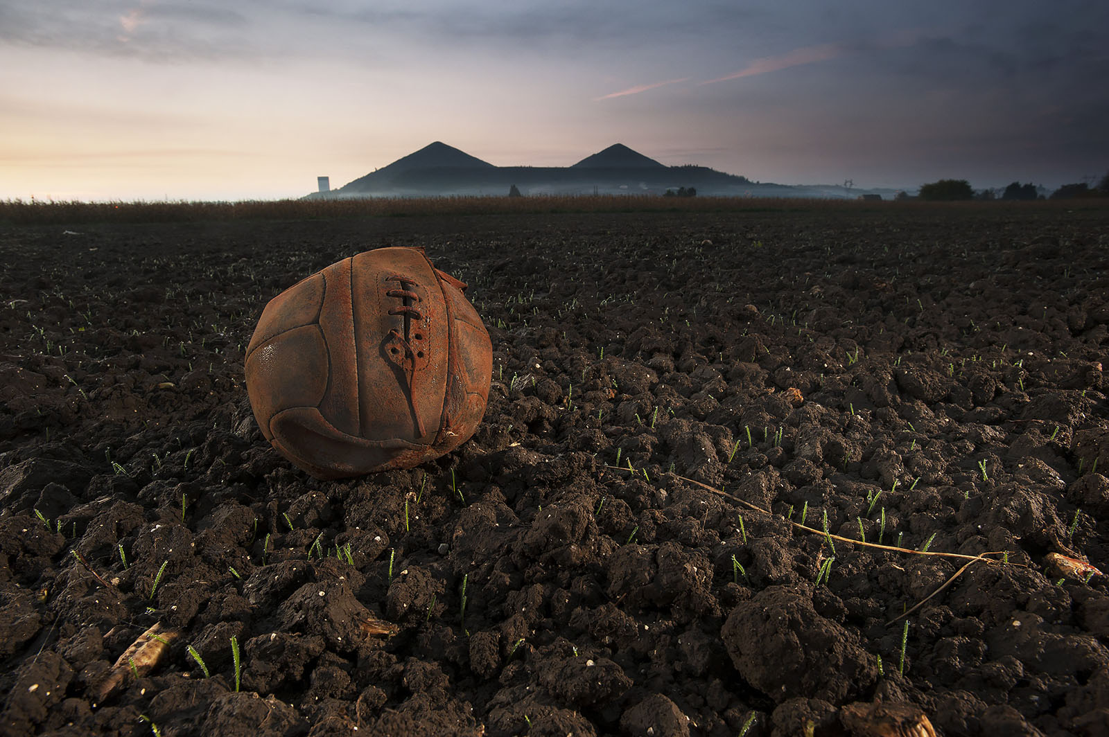 Modern photograph of a field of cracked dirt. A deflated brown football/soccer ball sits in the foreground.