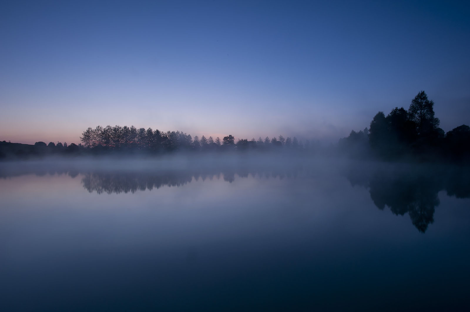 Modern photograph of morning mist rising off a glass-like water surface with trees in the background.