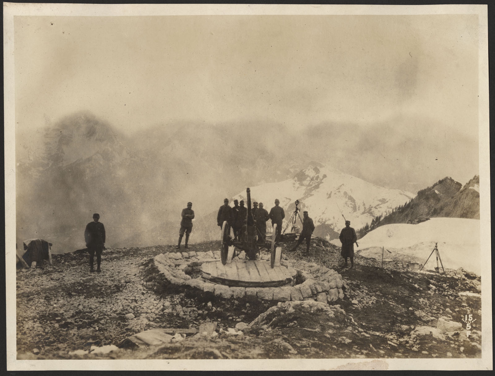 Vintage black and white photograph. A group of men stand behind a large gun on wheels in the mountains.