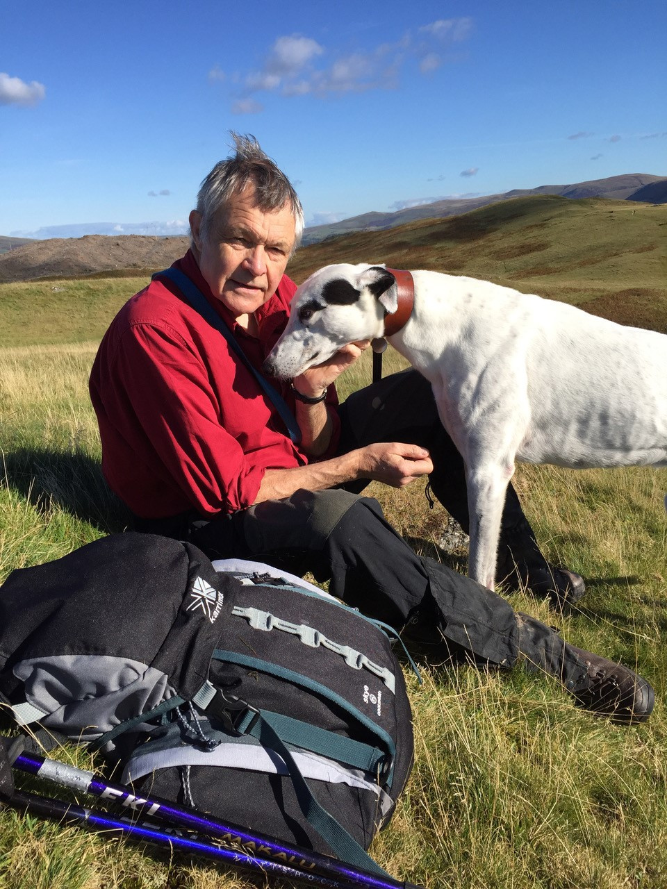 Modern photograph of an older white man wearing a red shirt sitting in a field next to a black and white dog.